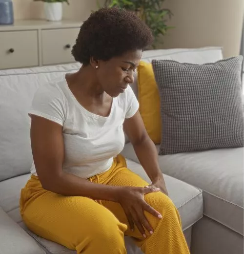 Black woman sitting on a couch, holding her knee and appearing in pain, representing joint discomfort caused by gout