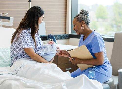 Woman in hospital bed holding newborn baby while sitting nurse looks at baby