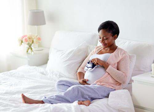 Black pregnant woman sitting up in bed placing baby shoes on her belly
