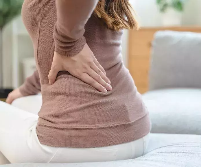 A person sitting on a couch, holding their lower back with one hand, likely experiencing back pain.